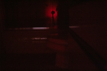 a camera on a tripod, backlit in red, reflected in a shower door beyond a toilet; a whitish-red glow is visible from under the door behind the camera