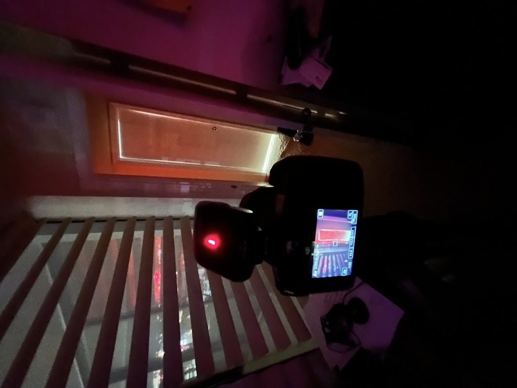 a camera in a dark room pointing at a door, beyond which is a brightly lit room; on the camera's rear LCD is the door