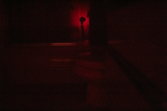 a camera on a tripod, backlit in red, reflected in a shower door beyond a toilet; a dim red glow is visible from under the door behind the camera