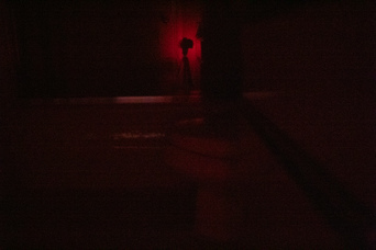 a camera on a tripod, backlit in red, reflected in a shower door beyond a toilet; a red glow with a hint of white is visible from under the door behind the camera